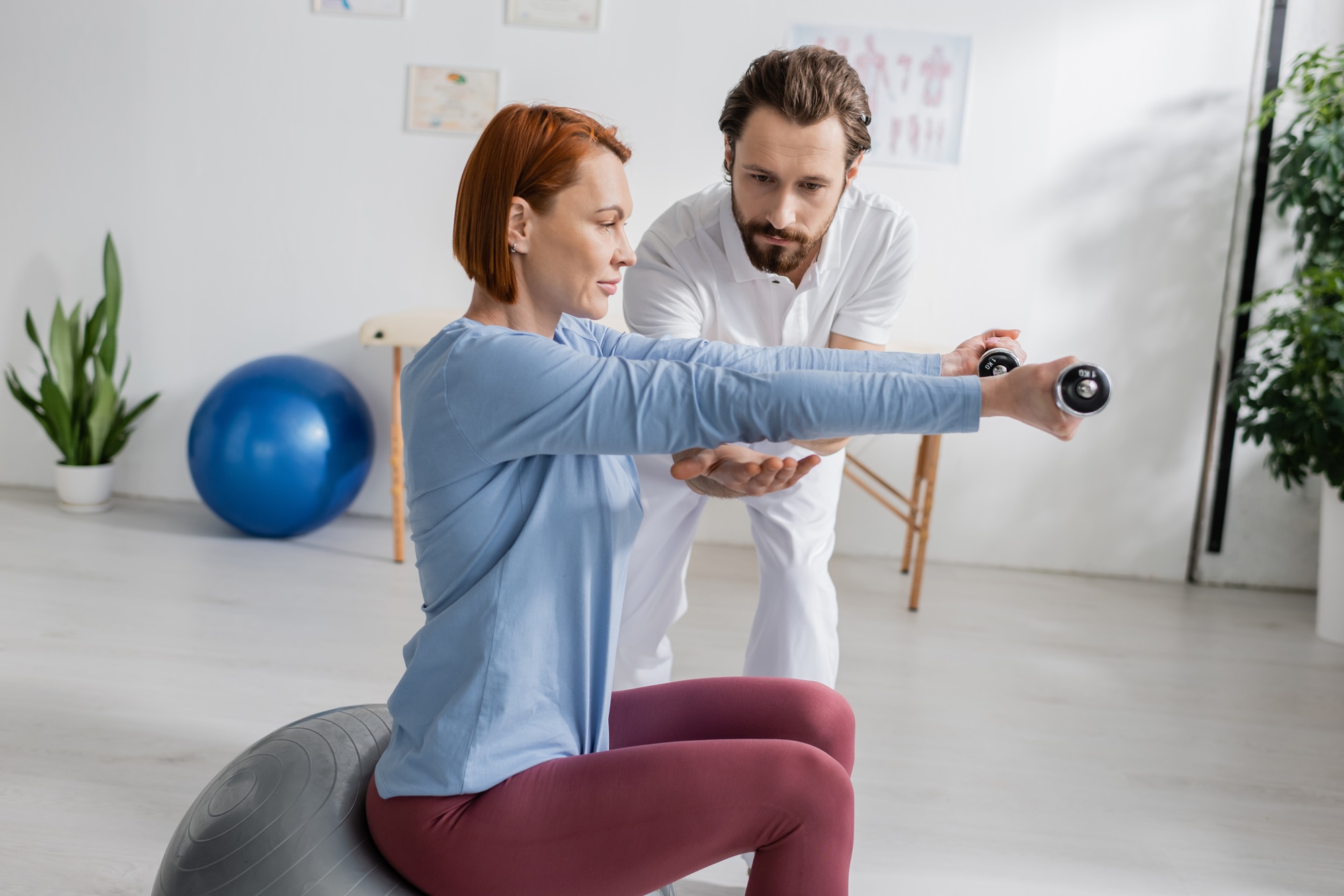 bearded physiotherapist assisting redhead woman sitting on fitball and training with dumbbells